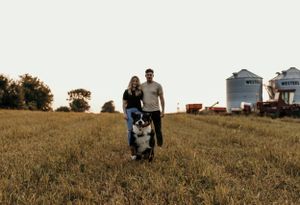 Couple walking with their dog through golden field at sunset with farm silos in background.