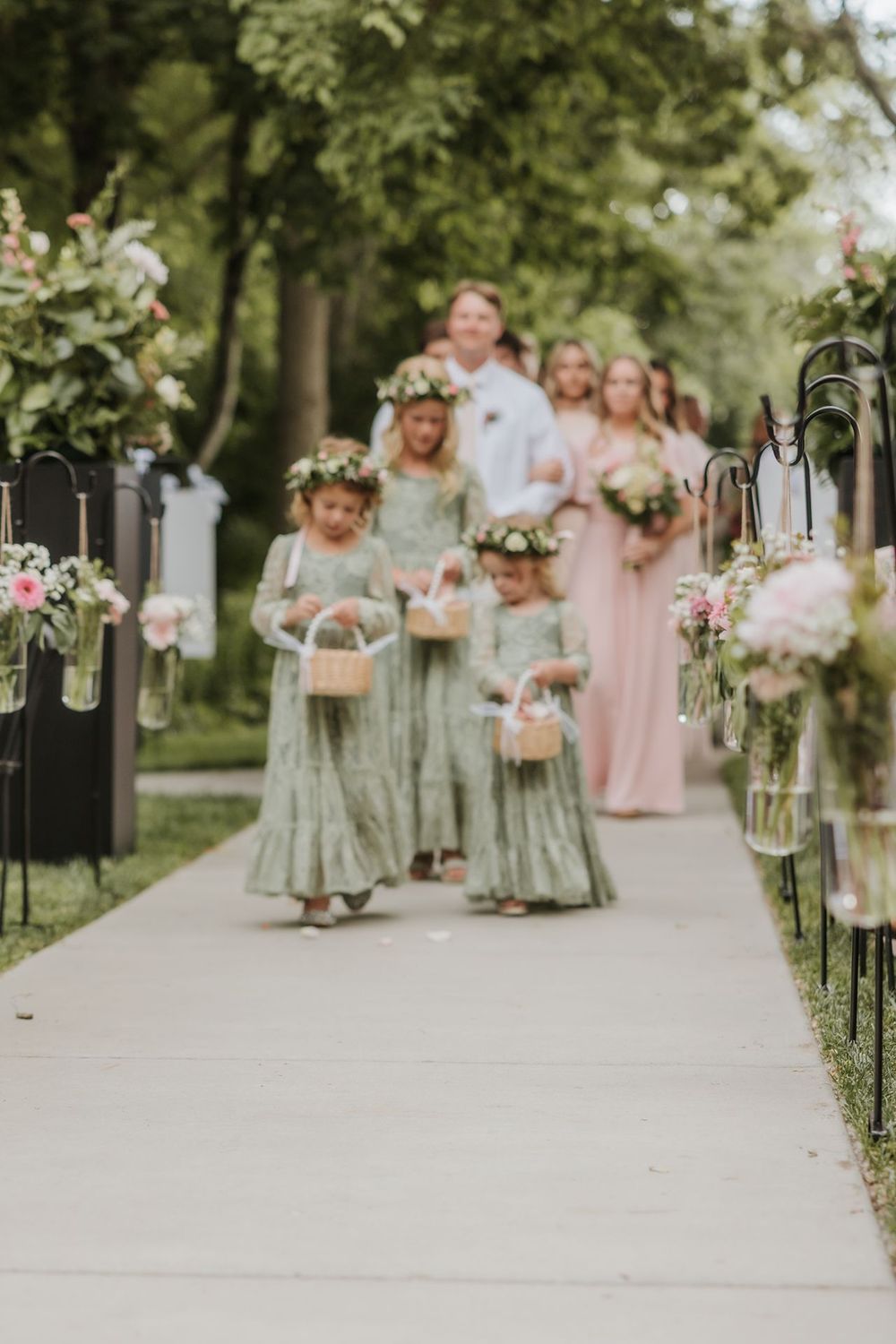 Wedding party in soft pastel dresses walks along tree-lined path with hanging lanterns.