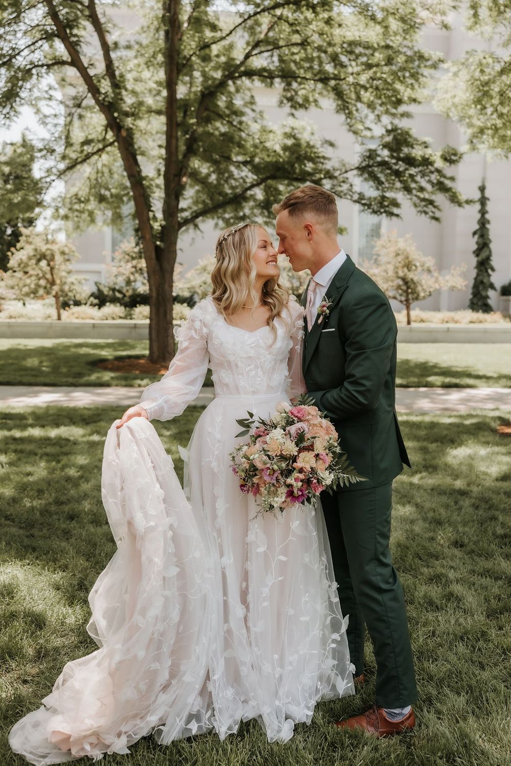 Couple shares intimate moment on sunny lawn with bride in detailed lace gown at LDS Timapnogos temple.