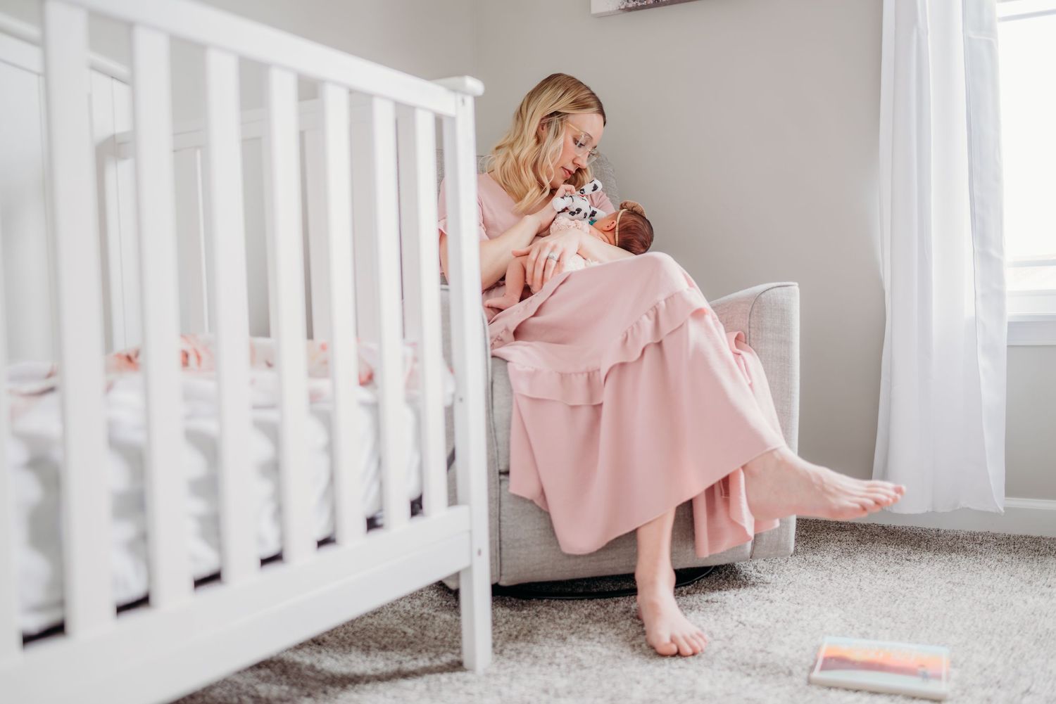 Parent in pink dress sits beside white crib holding sleeping newborn in soft nursery setting.