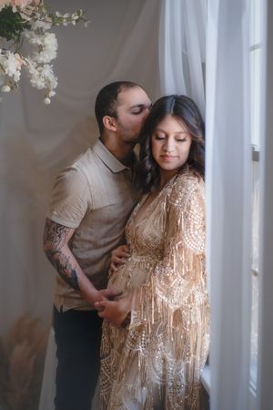 A romantic maternity photo of a couple embracing by a window with flowing white curtains and flowers.