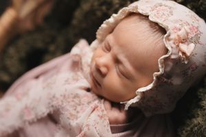 Sleeping newborn baby wearing vintage style pink lace bonnet and dress lying on dark textured blanket.