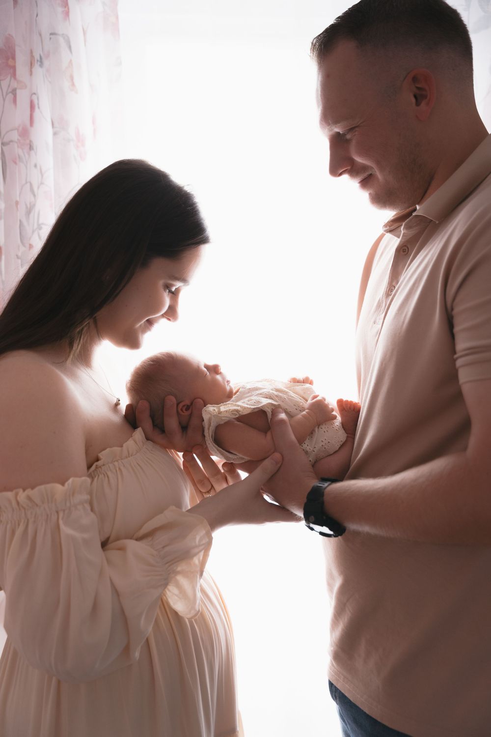 A tender family moment with a newborn baby in a sunlit room with pink curtains.