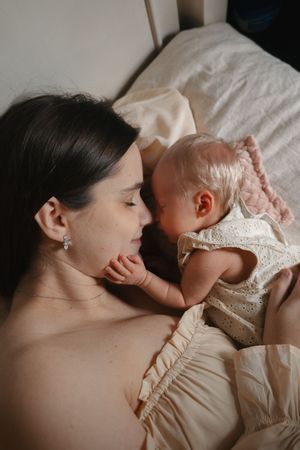 Mother and baby share intimate bonding moment while lying together on white bedding.