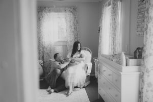 A black and white photo of someone sitting in a sunlit bedroom while being helped into a formal white dress.
