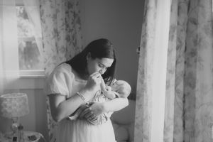 Black and white photo of woman in white dress holding and feeding newborn baby near window with curtains.