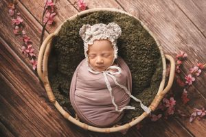 Swaddled newborn wearing a delicate knit bonnet rests in round basket with moss bedding and pink floral accents.