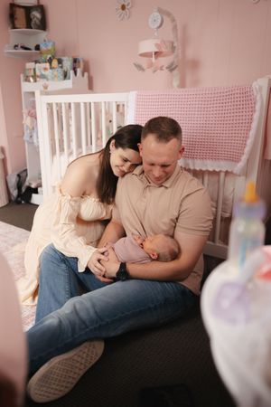 A tender moment between a couple sitting together in a pink nursery room with a white crib and knit blanket.