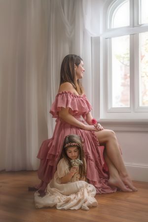 A mother and young daughter in pink and white dresses sitting by a sunlit window in a dreamy vintage-style portrait.