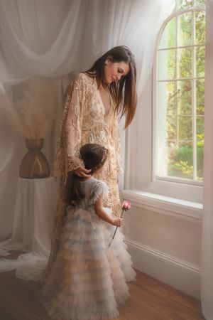 A dreamy portrait of two figures in lace and tulle dresses standing by a sunlit window with flowing white curtains.