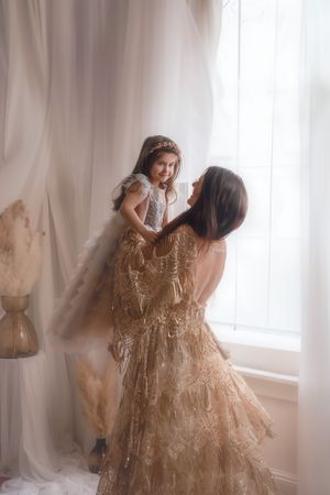 A mother and daughter share a tender moment by a window, both wearing elegant beige lace dresses in soft lighting.