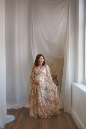 Elegant maternity portrait of woman in champagne lace gown against white backdrop.
