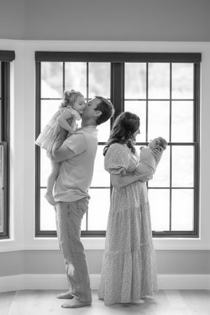 A black and white family portrait showing parents holding babies by a bright window.