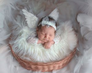 Sleeping newborn in an angel costume lies peacefully on white fluffy fur in a woven basket.