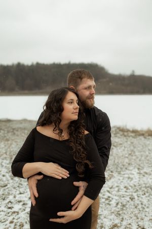 A couple in black clothing embraces on a snowy winter landscape with a lake and trees in the background.