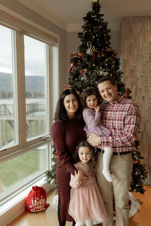 A family poses together near decorated Christmas trees and large windows overlooking a scenic winter landscape.
