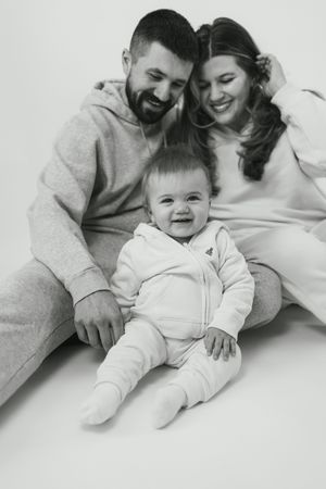 A black and white photo series shows tender moments between parents and their baby lying together on white bedding.