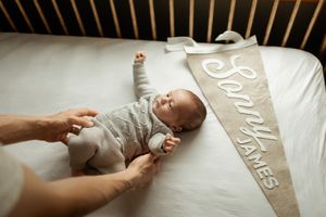 A small baby wearing a gray onesie lies on a white bed next to a decorative banner.