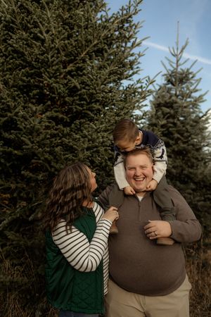 Two people share a candid moment of laughter while standing in front of evergreen trees.