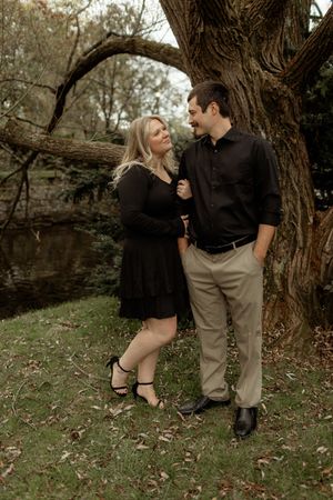 A couple in dark clothing poses together near a tree trunk in a romantic outdoor autumn setting.