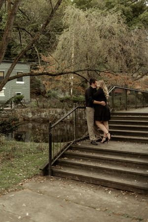 A couple embraces on stone steps in a garden setting with a weeping willow tree and rustic railing in the background.