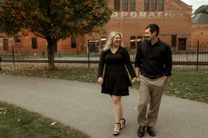 A couple walks hand in hand along a path on an autumn day with brick buildings and fall foliage in the background.