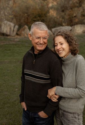 A mature couple wearing cozy sweaters embrace and smile warmly outdoors during an autumn photoshoot.