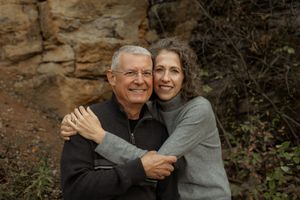 A couple embraces while smiling outdoors against a rocky cliff backdrop on an autumn day.