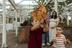 A person holds up autumn leaves while posing in front of a greenhouse or conservatory structure.