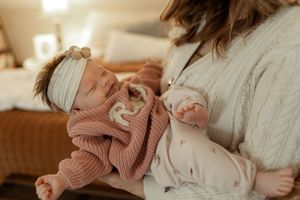 A sleeping newborn in a pink knit sweater and white headband rests against someone wearing a cozy white cardigan.