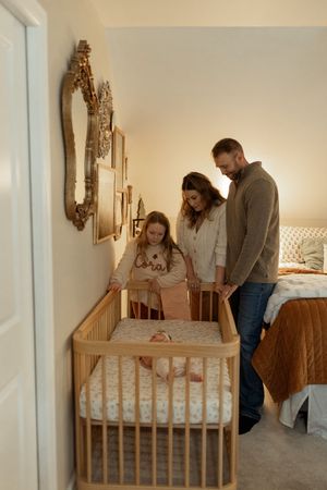Three progressive images show people setting up a wooden crib in a cozy bedroom with warm lighting.
