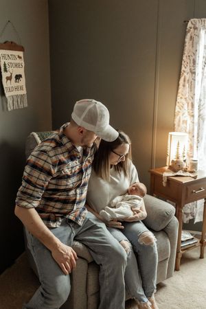 A couple sits on a gray couch in a cozy living room with rustic decor while holding a newborn baby.