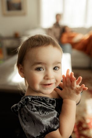 A young child waves at the camera while wearing a dark bandana around their neck in a warmly lit room.