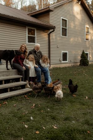 People gather outside a residential home to feed and interact with chickens on a grassy lawn during autumn.