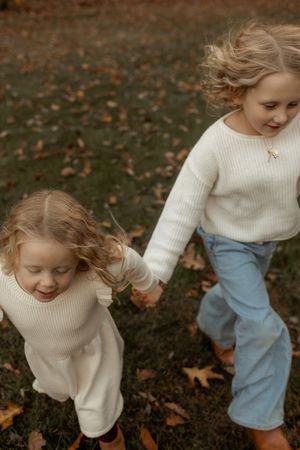 Two children in cream sweaters hold hands while playing in fallen autumn leaves on the ground.