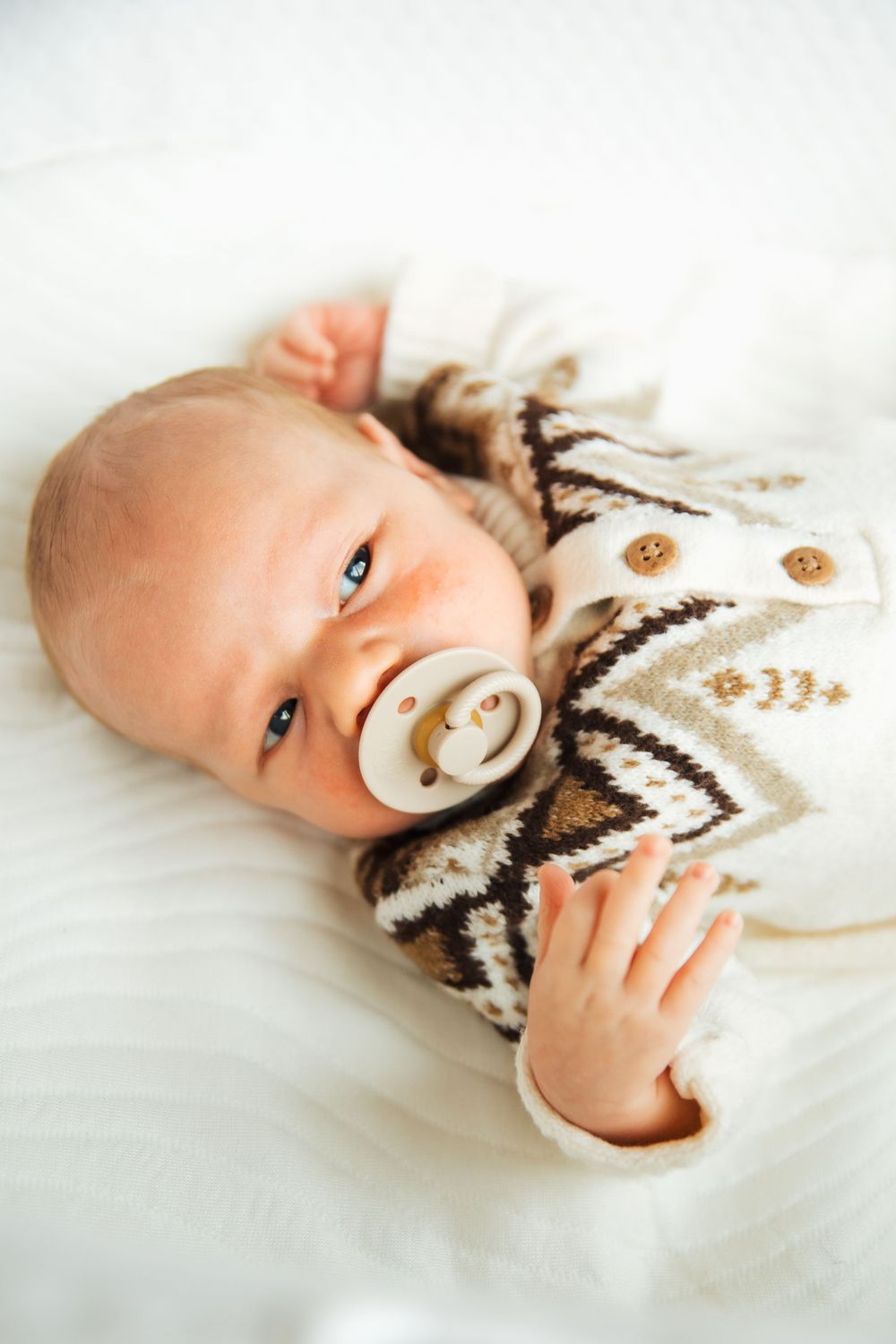Newborn baby with pacifier lying on white blanket wearing brown patterned knit onesie with buttons. Bristol Rhode Island Portsmouth