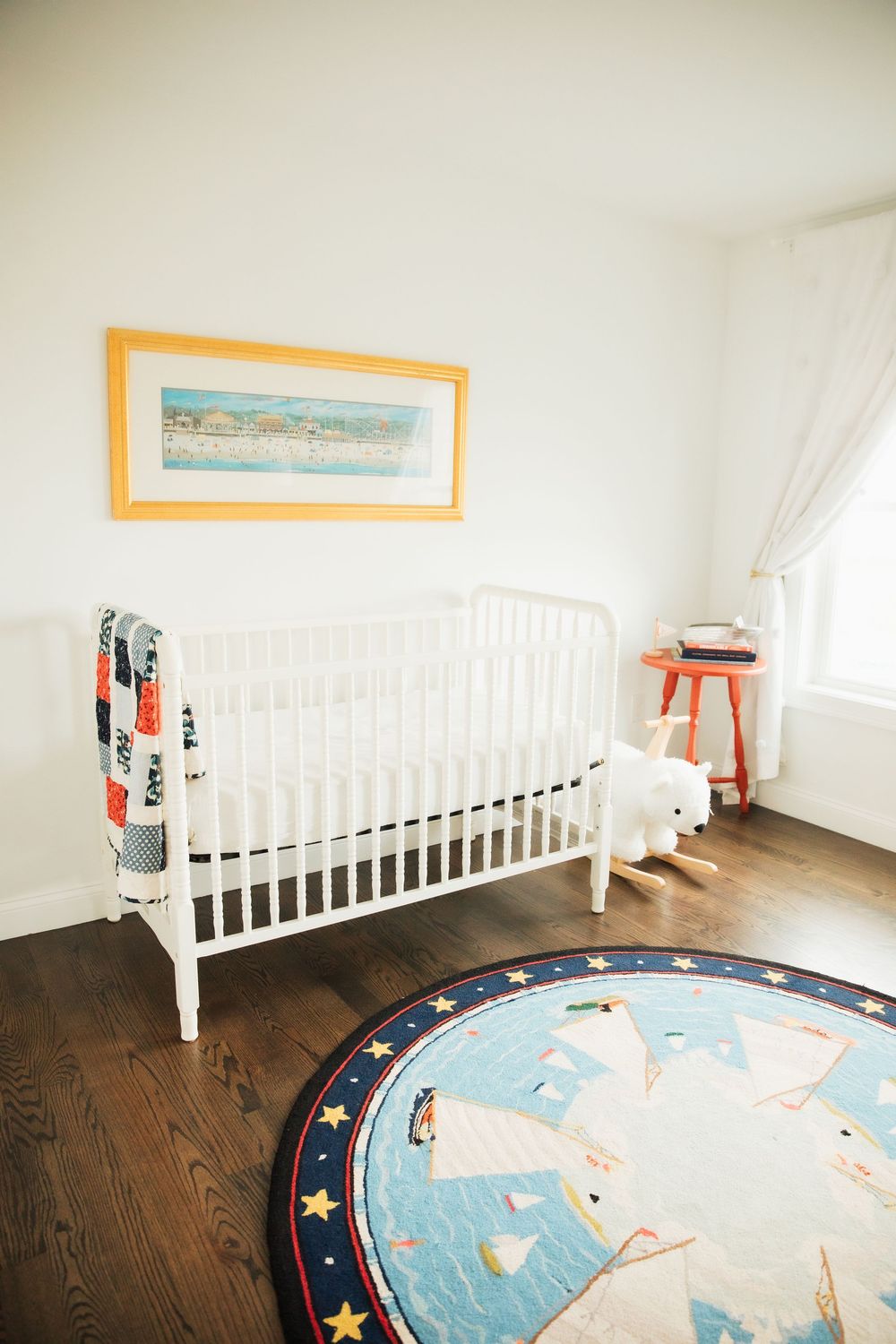 White baby crib in bright nursery with round unicorn rug, hardwood floors, and framed coastal artwork. Bristol Rhode Island Portsmouth