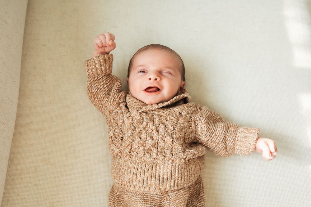 Smiling baby in a cozy brown knit sweater set lying on a cream surface with one arm raised. Bristol Rhode Island Portsmouth,