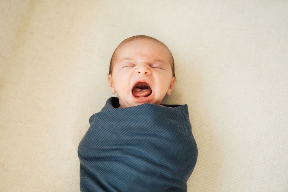 Newborn baby swaddled in a dark blue blanket yawning with eyes closed, lying on a beige background. Bristol Rhode Island Portsmouth,