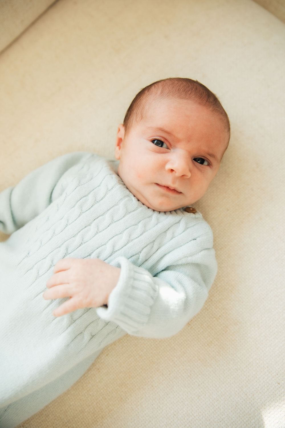 Newborn baby in light blue cable-knit onesie lying on cream surface, looking up at camera with alert eyes. Bristol Rhode Island Portsmouth,