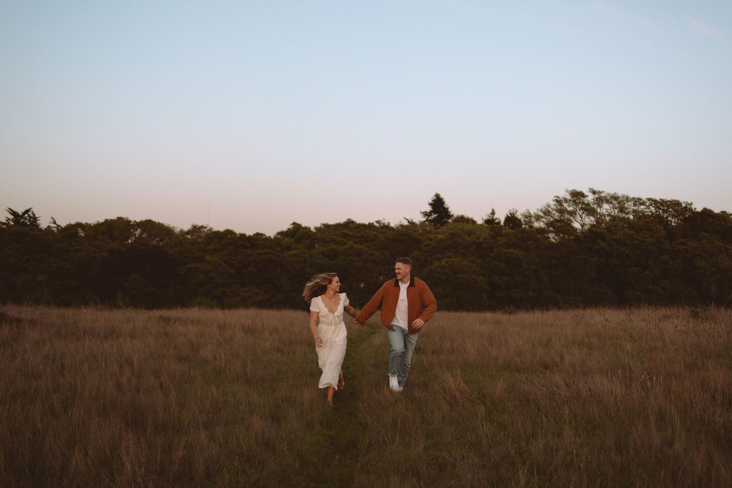 Two figures embrace in a golden field at dusk with trees silhouetted in the background.