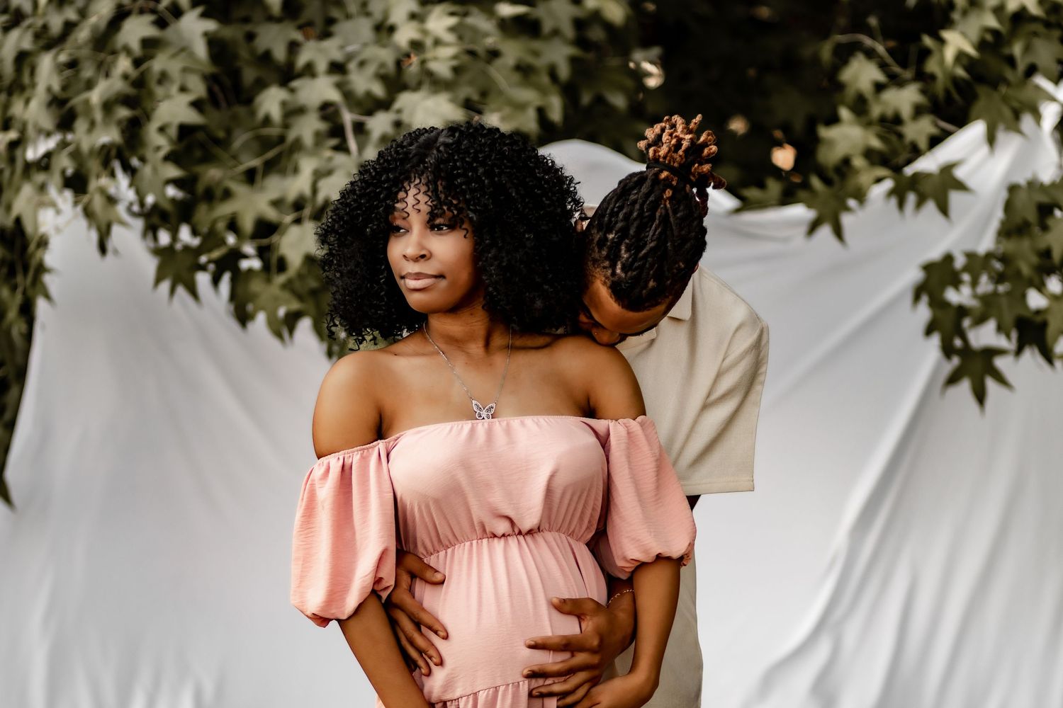 Expecting couple in a maternity photoshoot, woman in pink off-shoulder dress with partner embracing her baby bump.