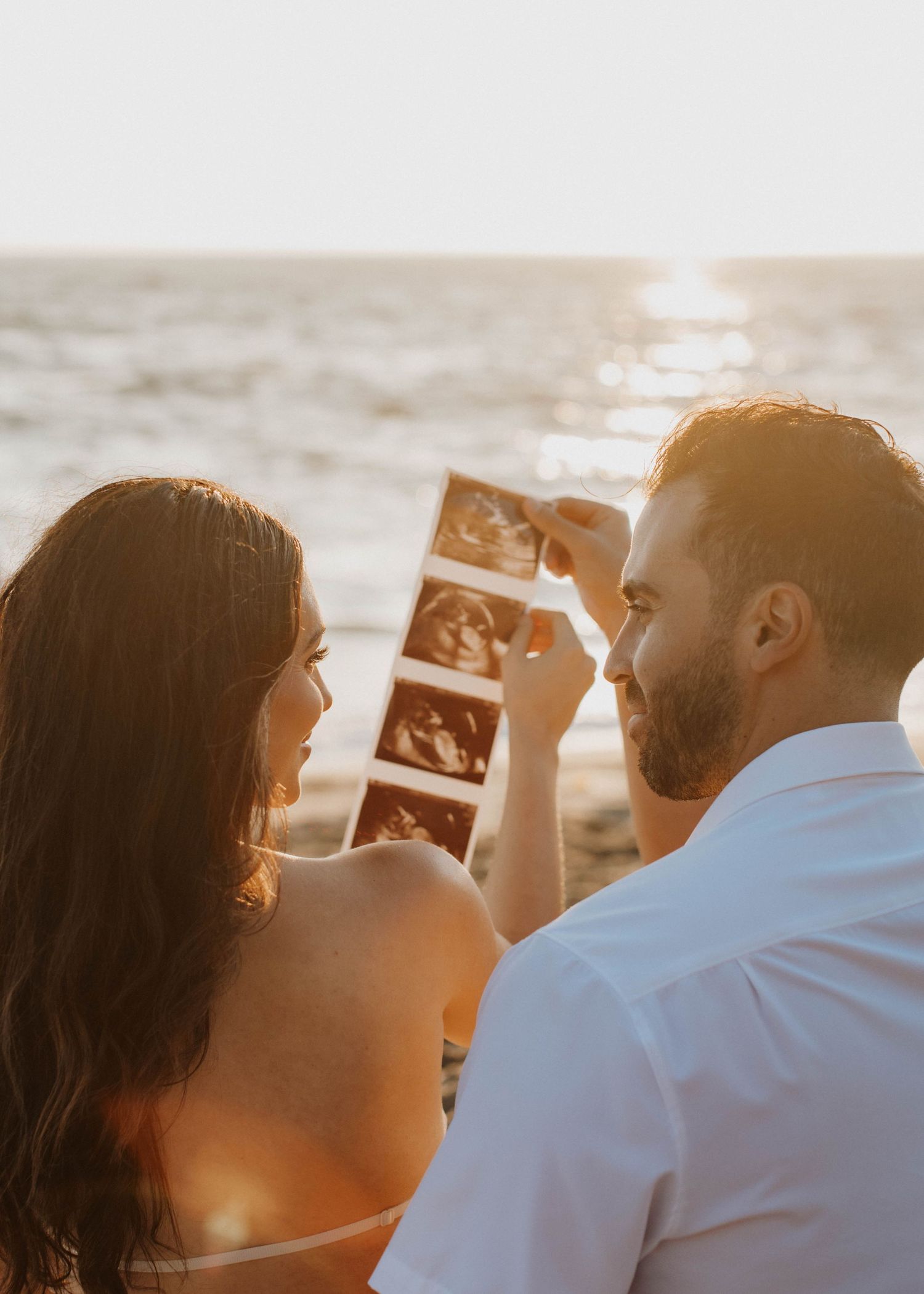 Couple holding ultrasound photos while sitting on beach at sunset.