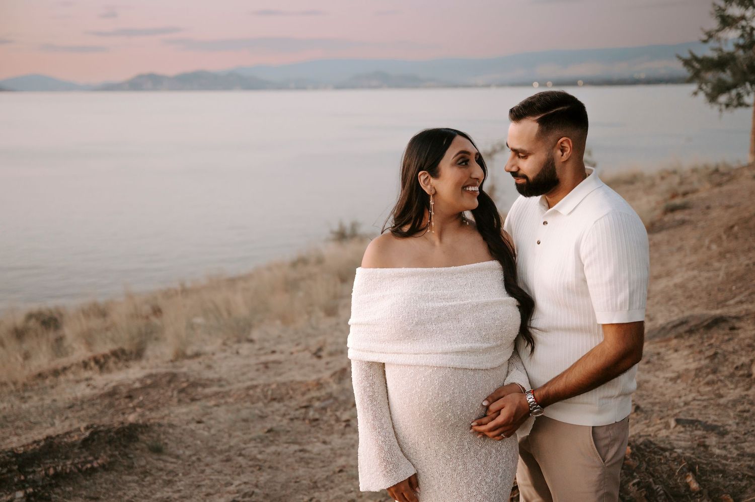 A pregnant woman in a flowing red dress stands on a scenic cliffside overlooking a lake at sunset.