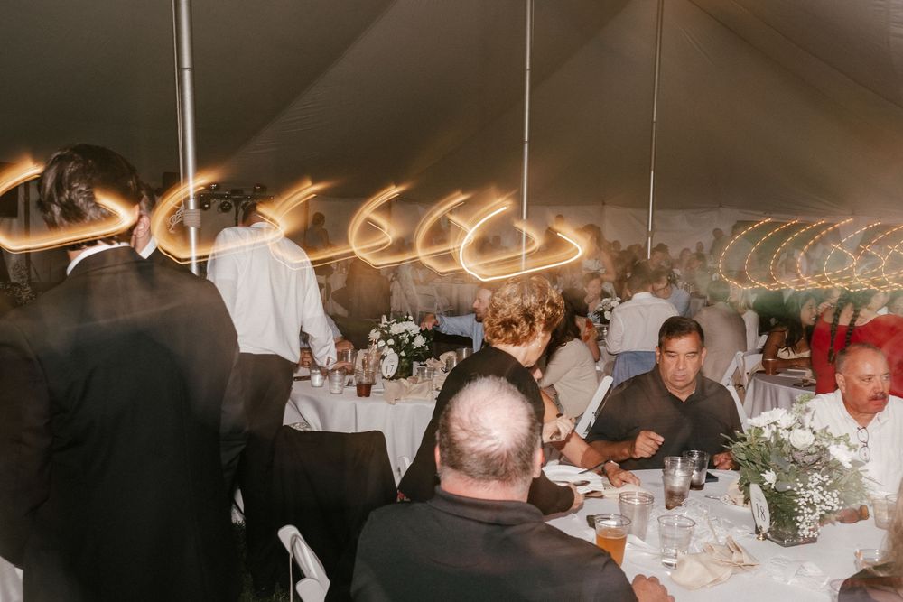 Guests dining under a white tent at an elegant wedding reception with warm glowing chandelier lights overhead.