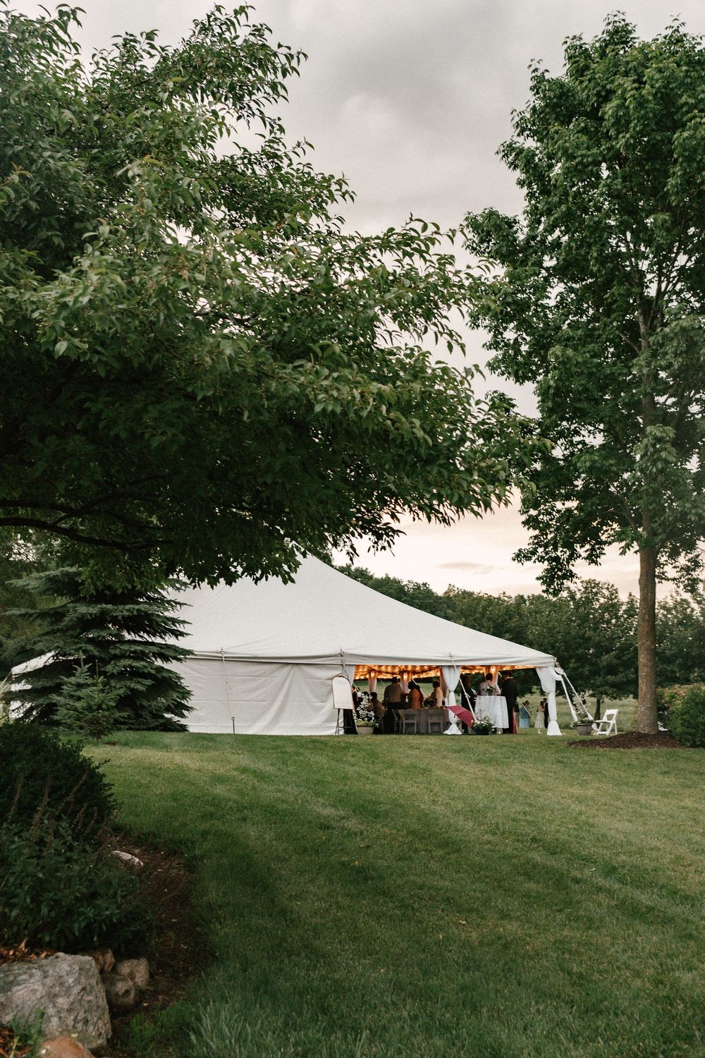 White event tent with string lights on lush green lawn, surrounded by trees under overcast sky.