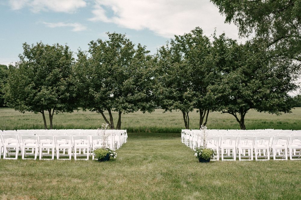 Outdoor wedding ceremony setup with white chairs arranged on green lawn beneath lush trees under a partly cloudy sky.