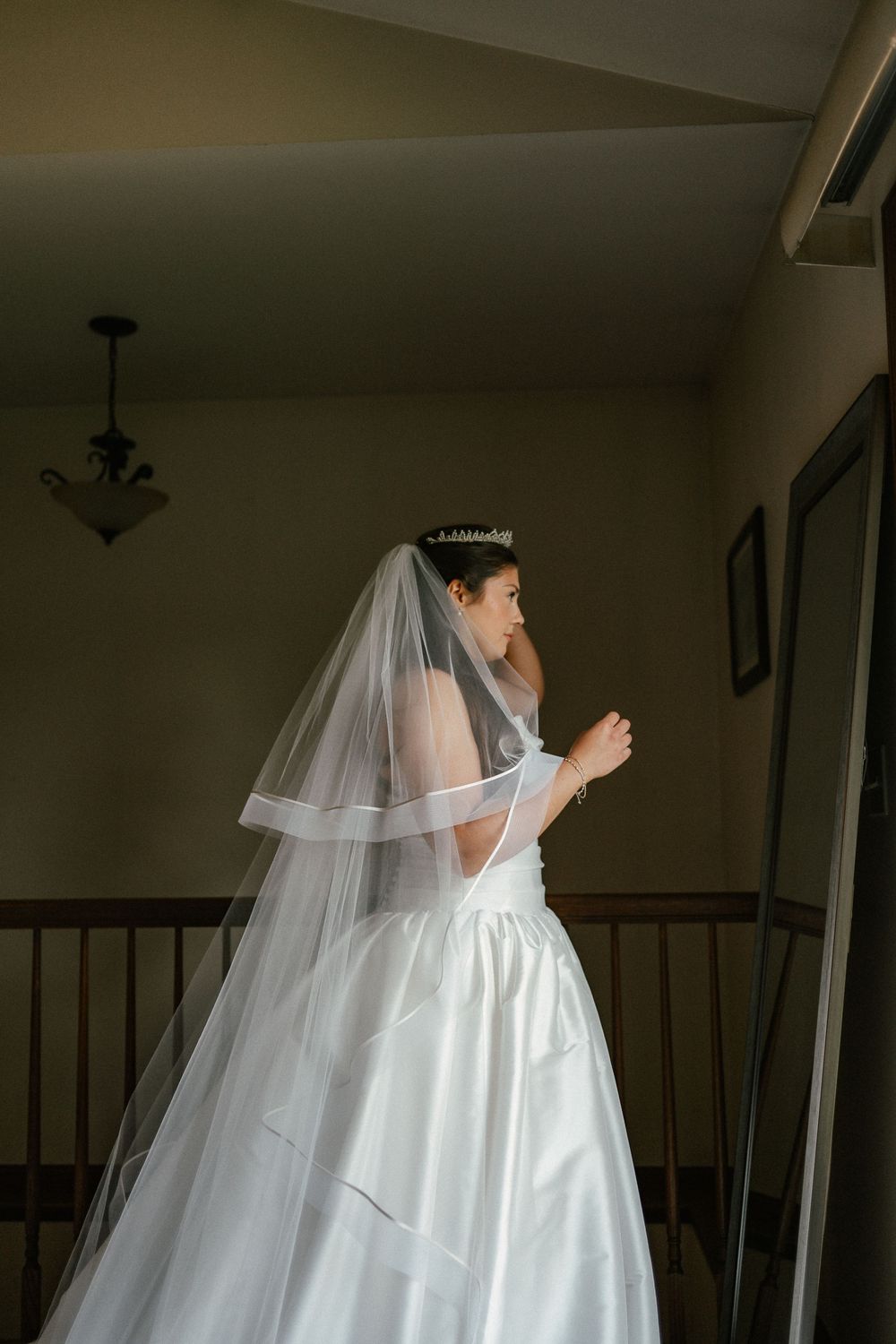 Bride in white satin gown and long veil with tiara stands near staircase railing in soft natural light.
