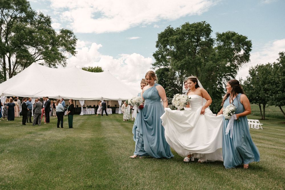 Bride in white gown walks with bridesmaids in blue dresses across green lawn near white wedding tent.