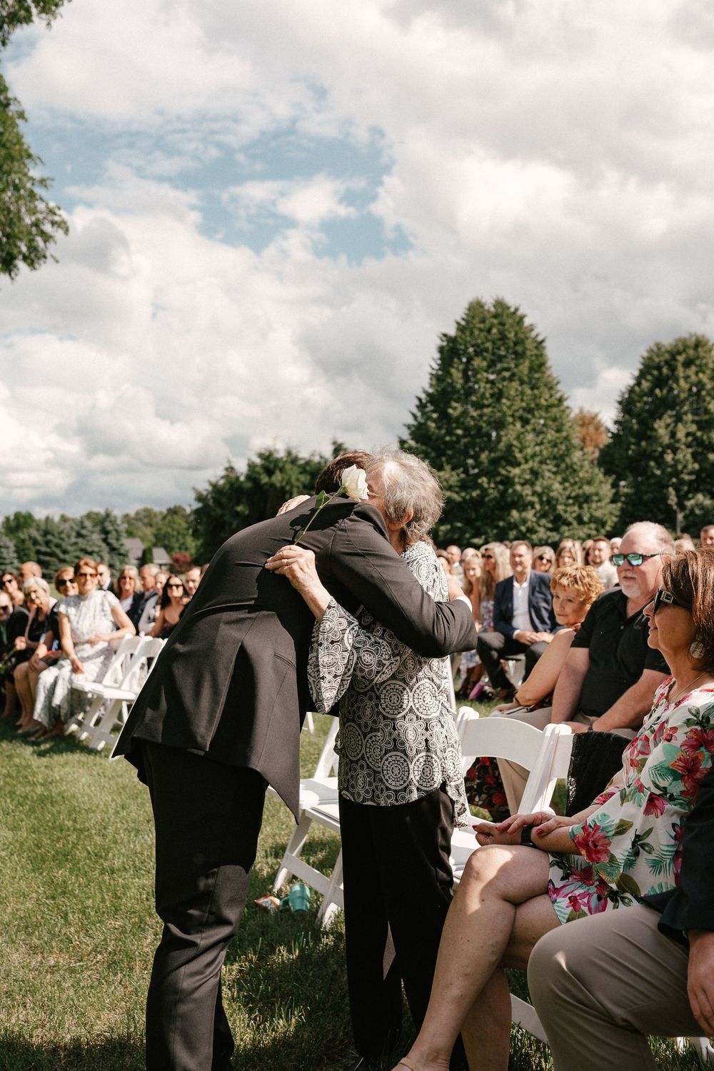 Two guests share an emotional hug during an outdoor wedding ceremony on a sunny day.